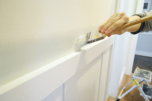 edging with a paintbrush to apply gray paint above a chair rail in hallway