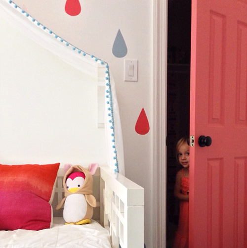 Garota espiando pela porta colorida do armário rosa ao lado de cama com mural de gotas de chuva | Cinco de Mayo