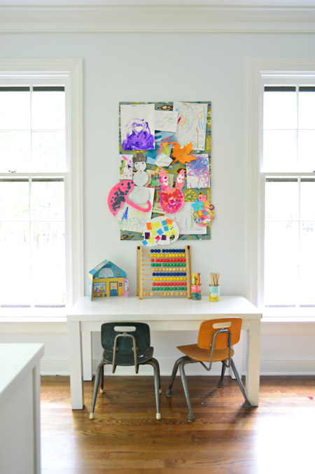 small fabric covered cork board used to pin up kid art projects above small homemade desk and schoolhouse style chairs