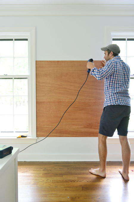 using power drill to screw plywood planks on wall between two windows to become base for gluing cork board tiles to create cork board wall